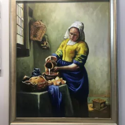 Reprodukcja obrazu 'Mleczarka' Vermeera w złotej ramie, przedstawiająca kobietę w stroju z epoki nalewającą mleko z dzbanka do miski.