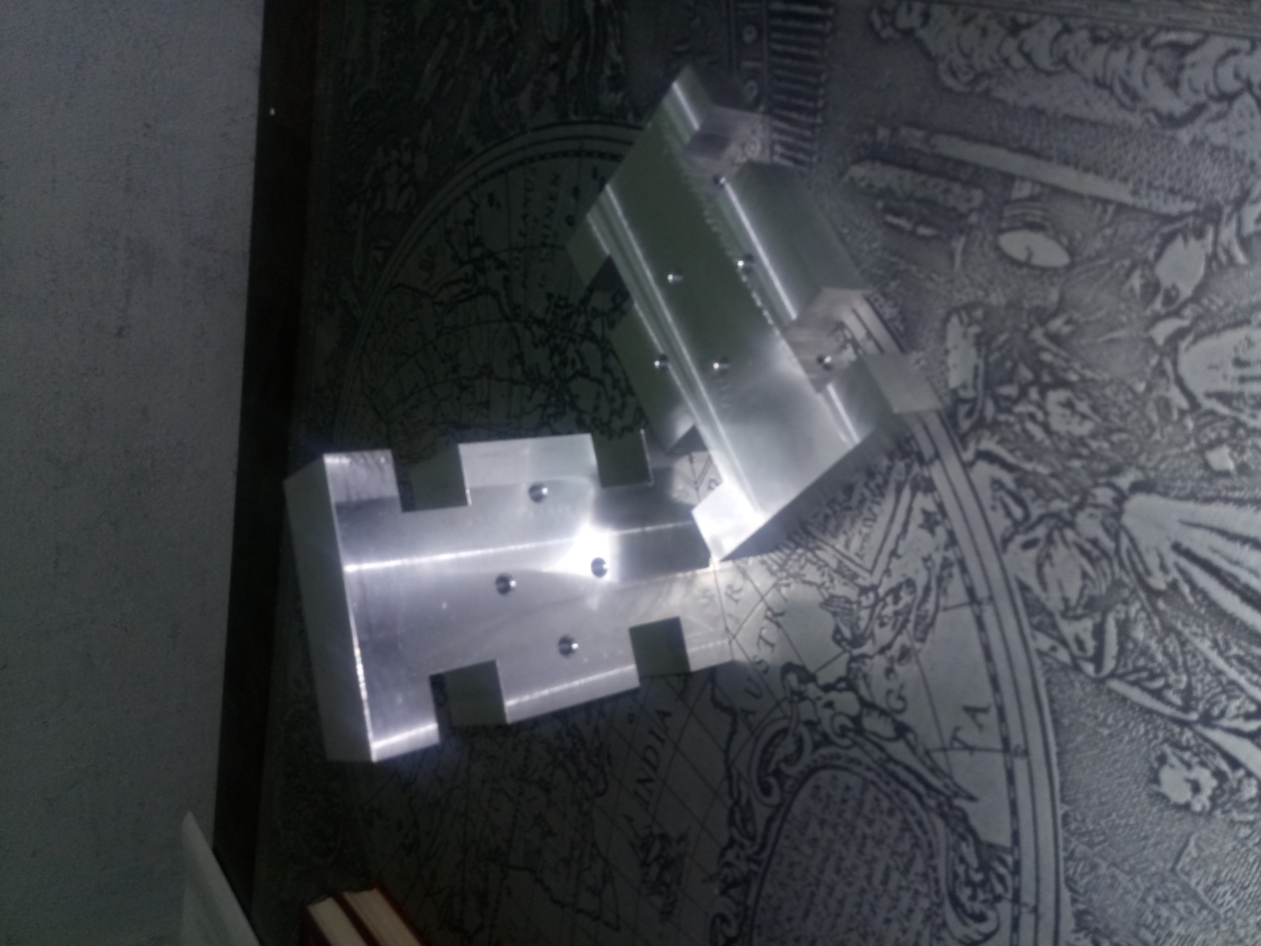 Dwa metalowe elementy obrobione CNC, leżące na tle mapy świata, widoczne ślady obróbki i otwory montażowe.