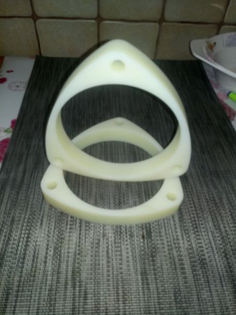 Dwa identyczne, białe, plastikowe elementy o trójkątnym kształcie z zaokrąglonymi rogami i otworem w każdym rogu, ułożone jeden na drugim na szarej, teksturowanej powierzchni.