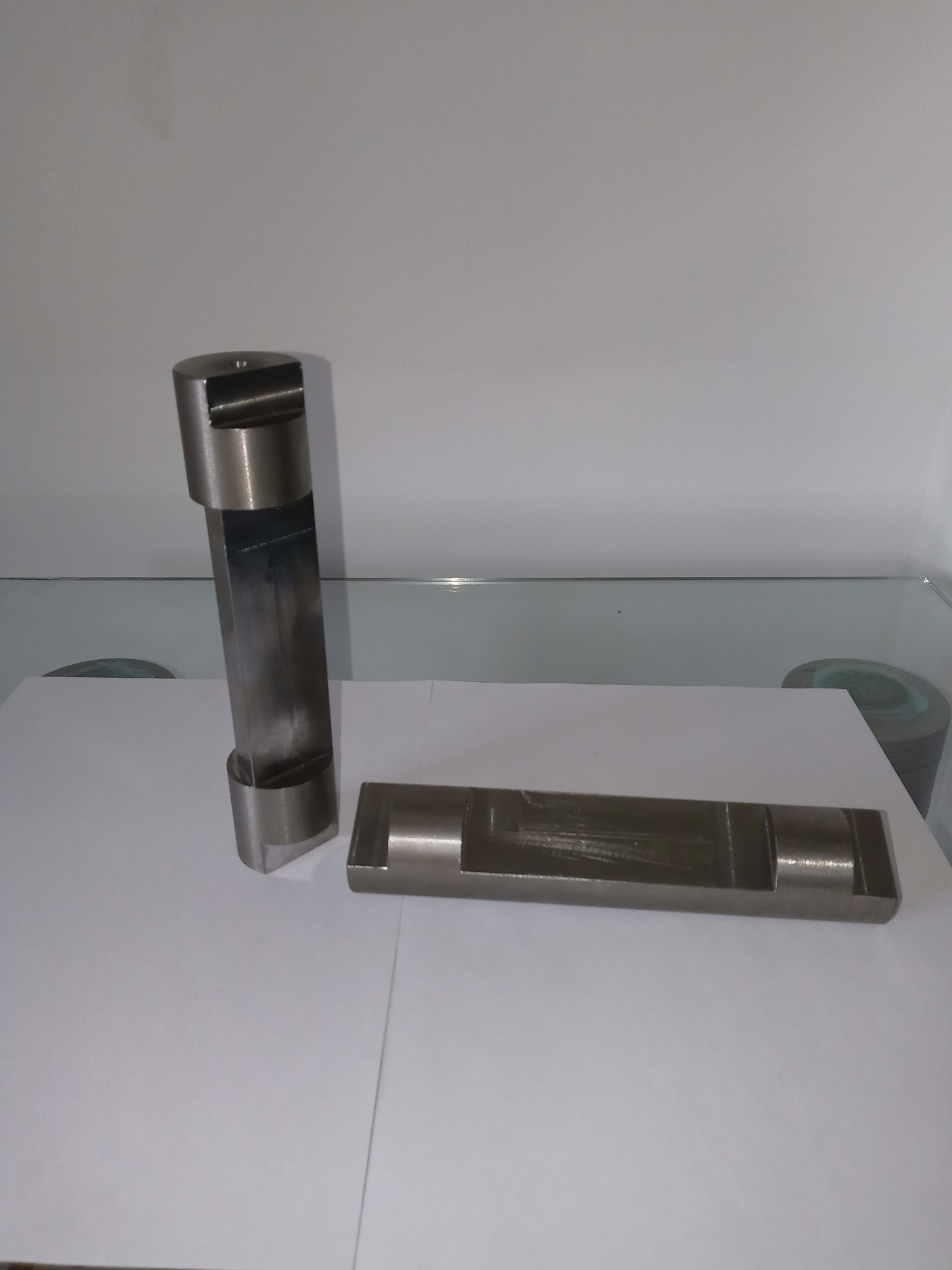 Dwa metalowe elementy obrobione maszynowo, jeden pionowy z wycięciem, drugi poziomy z wgłębieniem, leżące na białym tle.