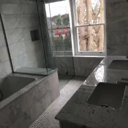 Wykończona łazienka z marmurowymi ścianami i podłogą w jodełkę; widok na wannę, kabinę prysznicową i dwie umywalki z bateriami; za oknem drzewa i budynki.