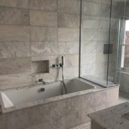 Cezary Pępczyński - Nowoczesna łazienka z wanną obudowaną marmurowymi płytkami, szklaną kabiną prysznicową i widokiem z okna na zabudowania.