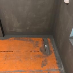 Cezary Pępczyński - Przygotowanie do hydroizolacji w kabinie prysznicowej: pomarańczowa mata drenażowa Schluter-DITRA 25 na podłodze, szare ściany z nałożoną warstwą hydroizolacji w narożniku pomieszczenia.