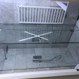 Cezary Pępczyński - Szklana kabina prysznicowa z widocznymi mocowaniami i taśmą zabezpieczającą na szkle, marmurowe płytki na ścianach i podłodze, zamontowana armatura prysznicowa.