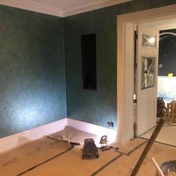 Cezary Pępczyński - Pokój w trakcie malowania ścian na zielono-niebieski kolor, widoczne lampy budowlane oświetlające ścianę, zabezpieczona podłoga i otwór w ścianie.