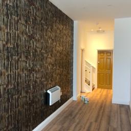 Cezary Pępczyński - Pokój po remoncie z tapetą o teksturze drewna na jednej ścianie, zamontowany grzejnik elektryczny i dwie puszki farby na podłodze z paneli.