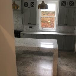 Cezary Pępczyński - Nowoczesna kuchnia z szarymi szafkami, marmurowym blatem i podłogą, widok zza framugi drzwi.