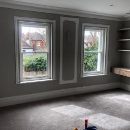 Cezary Pępczyński - Pokój z dwoma oknami w białych ramach, szarymi ścianami z dekoracyjnymi listwami, szarą wykładziną i drewnianą półką. Widoczne narzędzia malarskie na podłodze sugerują świeżo zakończony remont.