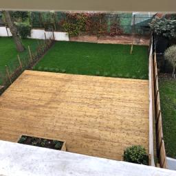 Cezary Pępczyński - Widok z góry na drewniany taras z wbudowanymi rabatami, przylegający do zadbanego trawnika z ozdobnymi nasadzeniami i ogrodzeniem.