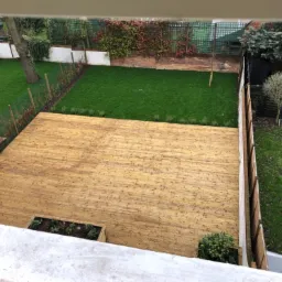 Widok z góry na drewniany taras z wbudowanymi rabatami, przylegający do zadbanego trawnika z ozdobnymi nasadzeniami i ogrodzeniem.