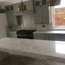 Cezary Pępczyński - Wykończona kuchnia z szarymi szafkami, marmurowym blatem i złotymi lampami wiszącymi, widok na ceglany budynek przez okno.