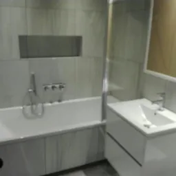 Nowoczesna łazienka z wanną obudowaną płytkami imitującymi marmur, bateria wannowa podtynkowa z trzema pokrętłami, umywalka nablatowa na szafce z dwiema szufladami i lustro w białej ramie.