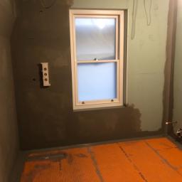 Cezary Pępczyński - Przygotowanie łazienki do położenia płytek: ściany zagruntowane, na podłodze pomarańczowa mata izolacyjna, widoczne okno i instalacja pod baterię.