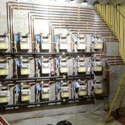 Cezary Pępczyński - Rząd liczników gazowych zamontowanych na ścianie z betonowych płyt, z rurami miedzianymi i żółtymi rurami gazowymi, drabina w tle.