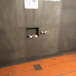 Przygotowana do wykończenia łazienka z pomarańczową matą grzewczą na podłodze, wnęką w ścianie i wyprowadzonymi instalacjami hydraulicznymi, na ścianie widoczny plan techniczny.