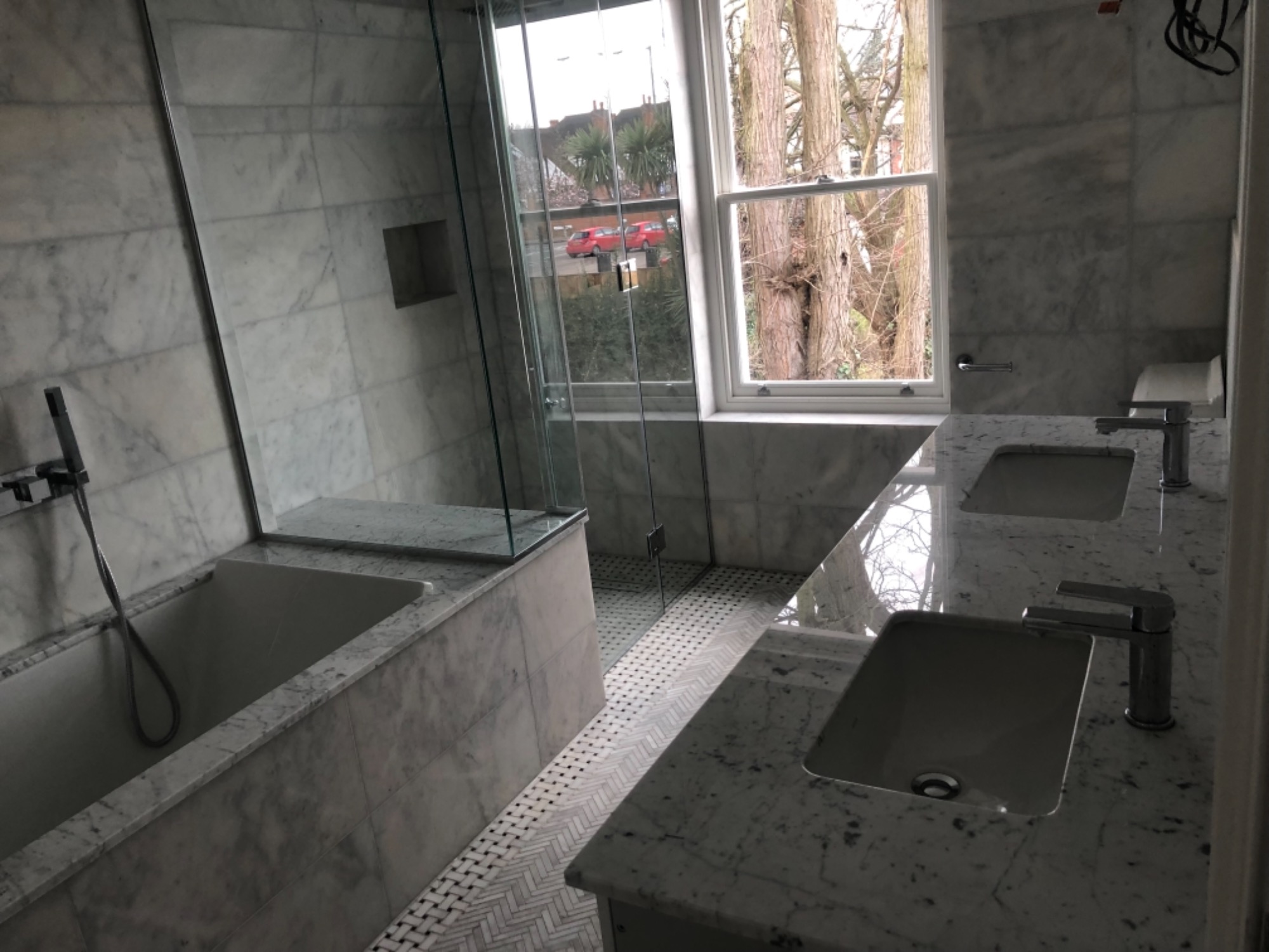 Wykończona łazienka z marmurowymi ścianami i podłogą w jodełkę; widok na wannę, kabinę prysznicową i dwie umywalki z bateriami; za oknem drzewa i budynki.