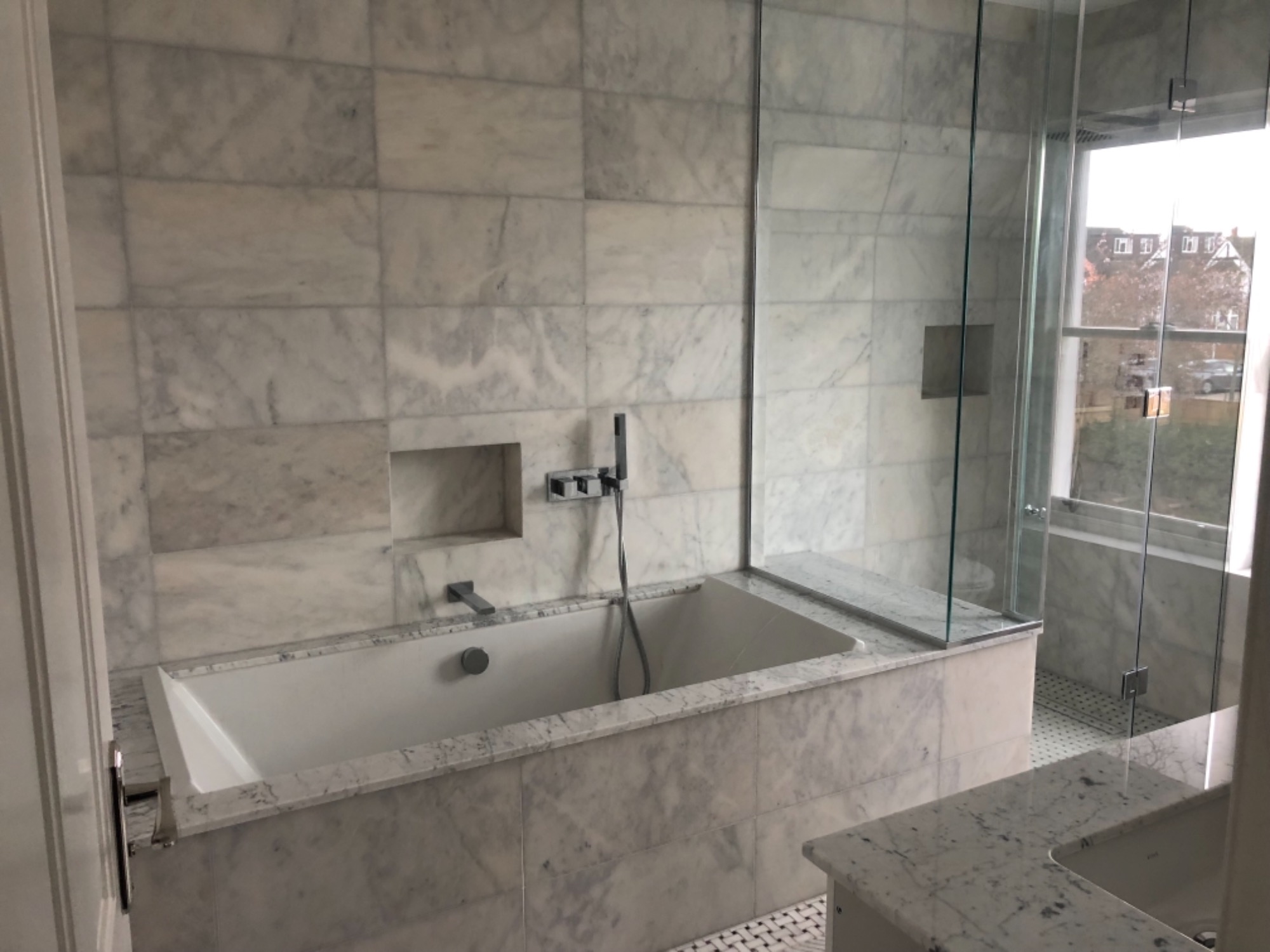 Nowoczesna łazienka z wanną obudowaną marmurowymi płytkami, szklaną kabiną prysznicową i widokiem z okna na zabudowania.