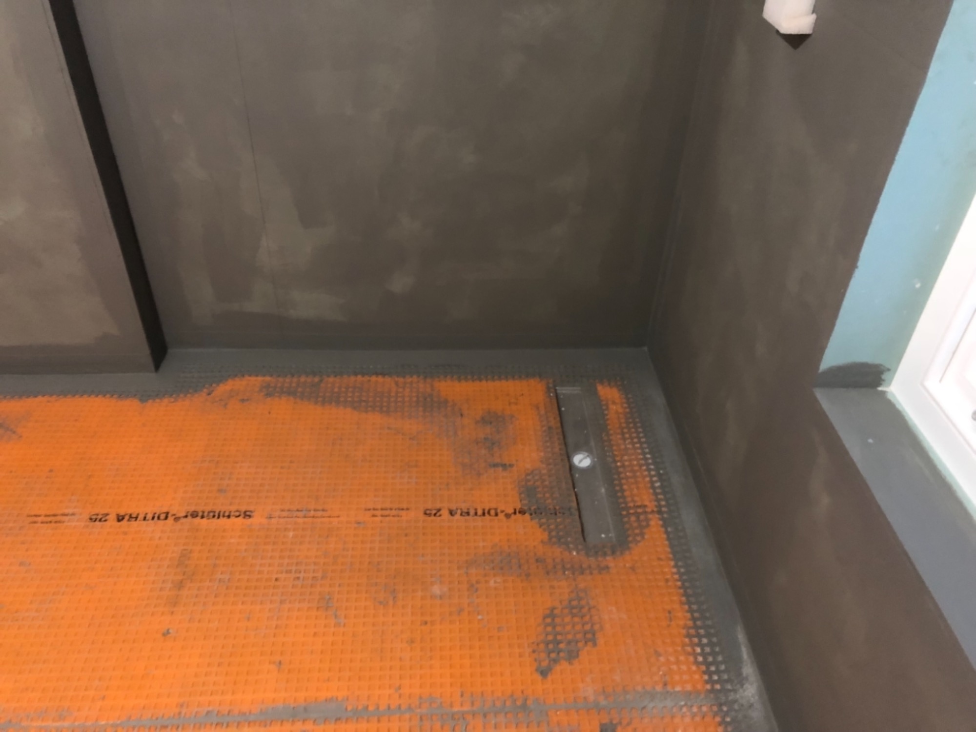 Przygotowanie do hydroizolacji w kabinie prysznicowej: pomarańczowa mata drenażowa Schluter-DITRA 25 na podłodze, szare ściany z nałożoną warstwą hydroizolacji w narożniku pomieszczenia.