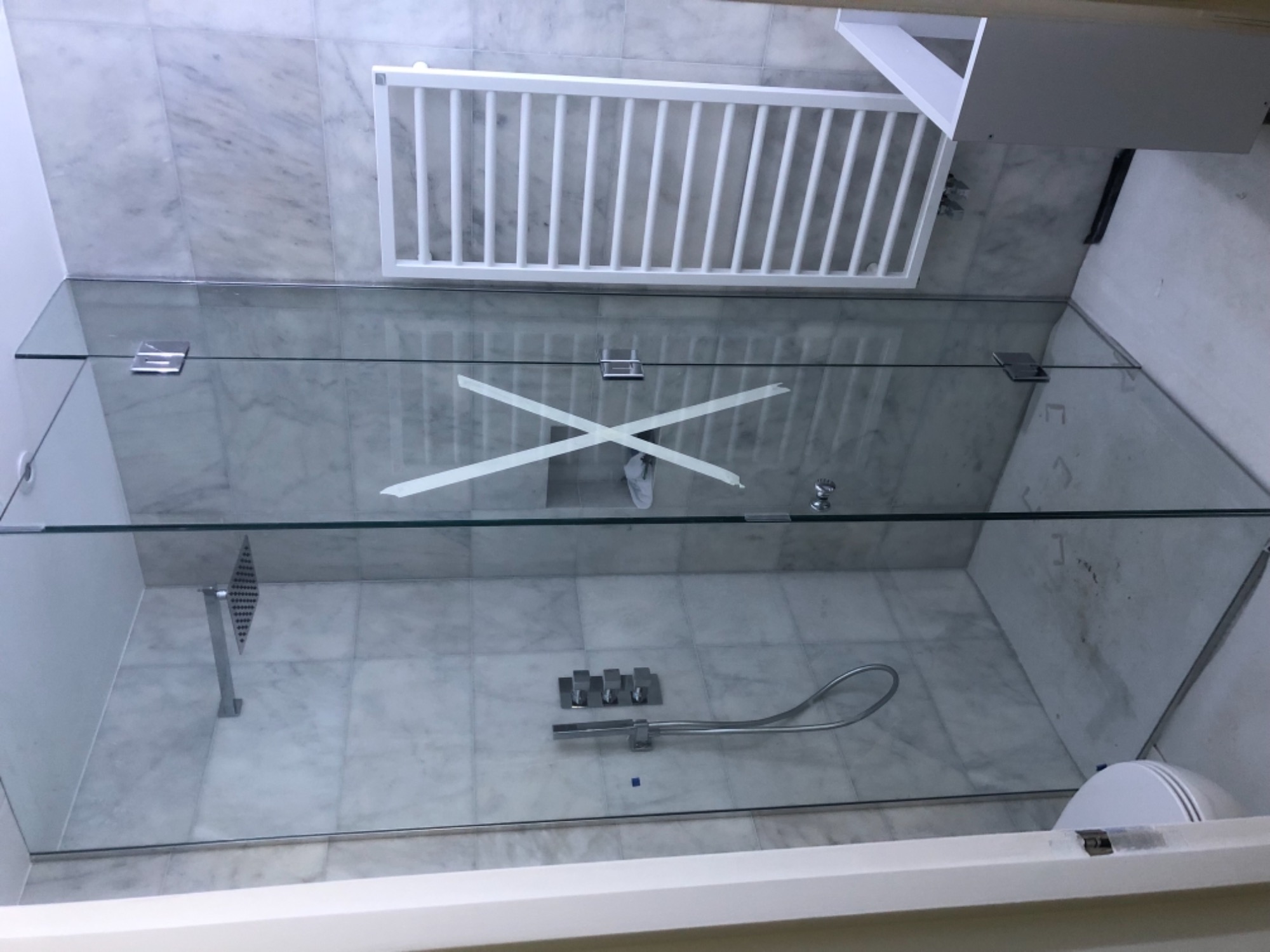 Szklana kabina prysznicowa z widocznymi mocowaniami i taśmą zabezpieczającą na szkle, marmurowe płytki na ścianach i podłodze, zamontowana armatura prysznicowa.