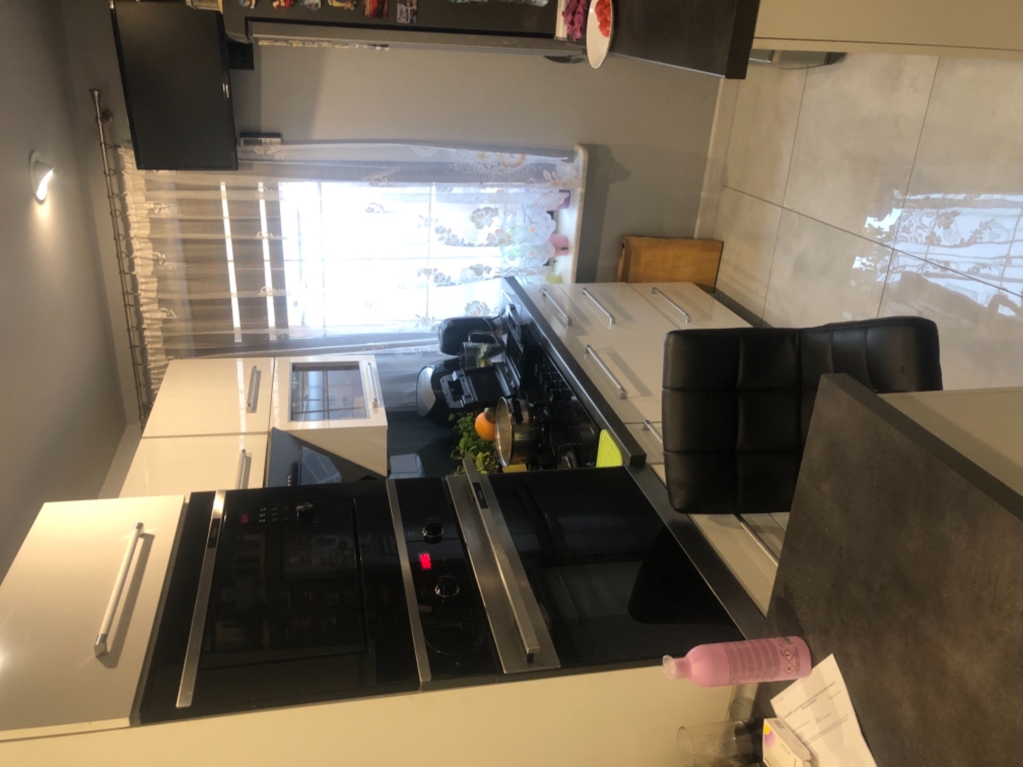 Nowoczesna kuchnia z zabudową meblową w odcieniach szarości i bieli, widoczny piekarnik, płyta grzewcza, krzesło barowe i okno z firanką.