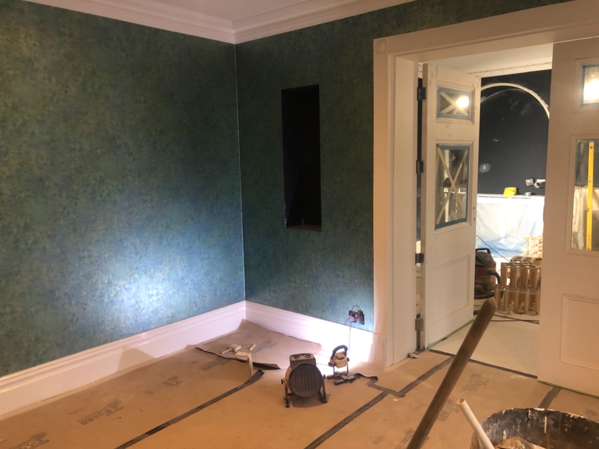 Pokój w trakcie malowania ścian na zielono-niebieski kolor, widoczne lampy budowlane oświetlające ścianę, zabezpieczona podłoga i otwór w ścianie.