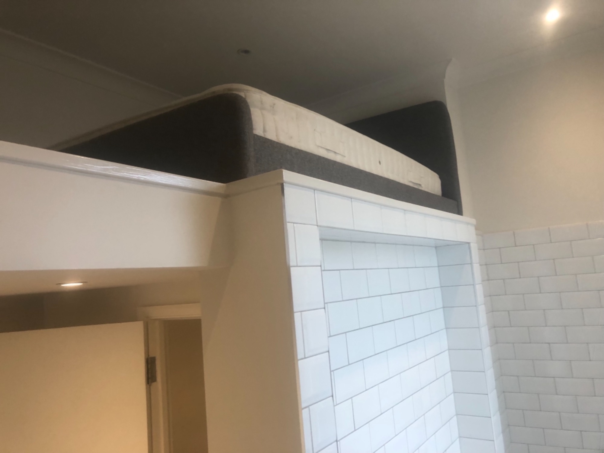 Materac na antresoli nad zabudowaną białą ścianą z płytek w łazience.