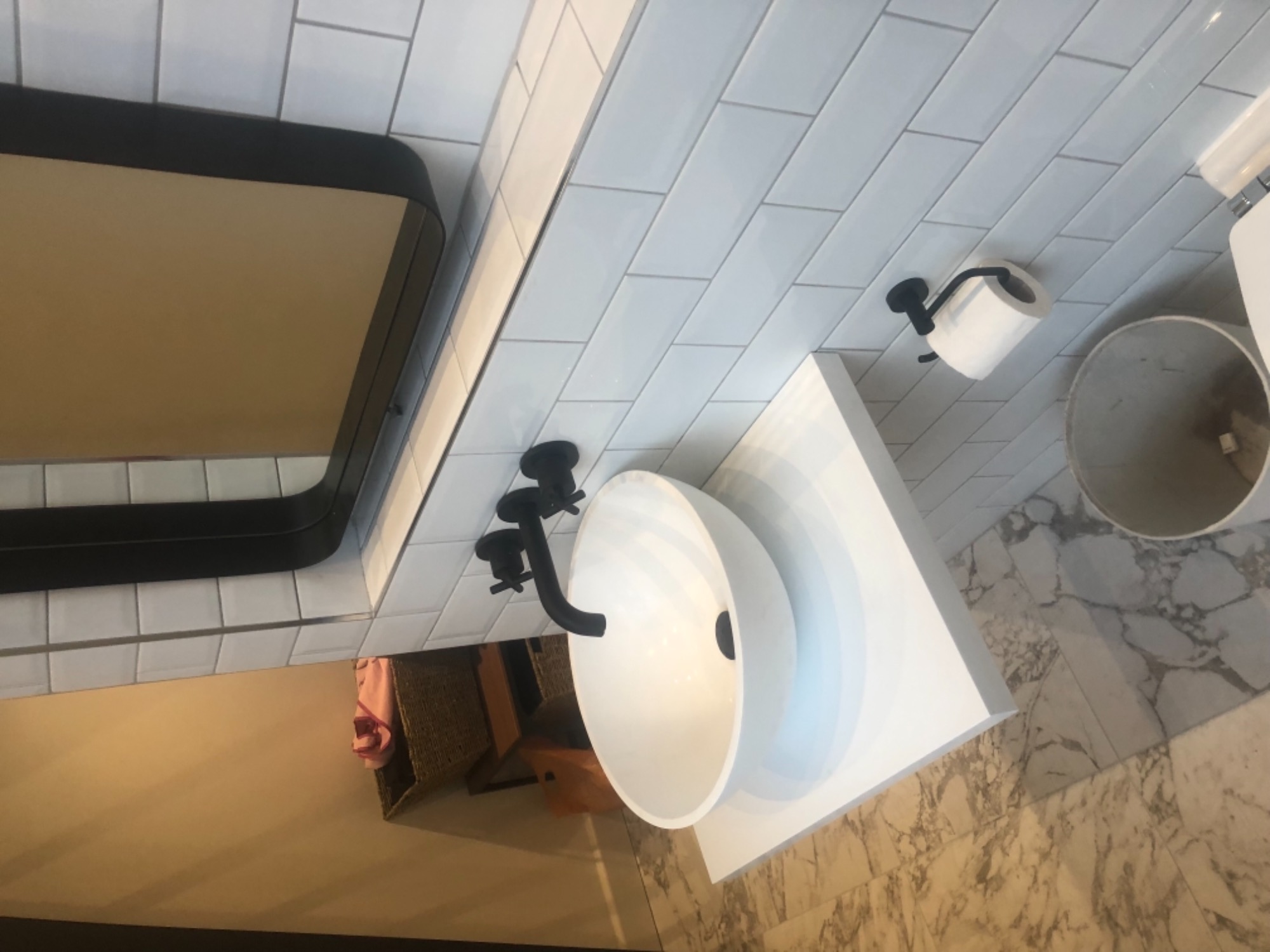 Nowoczesna łazienka z białą umywalką nablatową, czarną armaturą i dużym lustrem w czarnej ramie na tle białych, prostokątnych płytek ściennych, uzupełniona marmurową podłogą i akcesoriami...