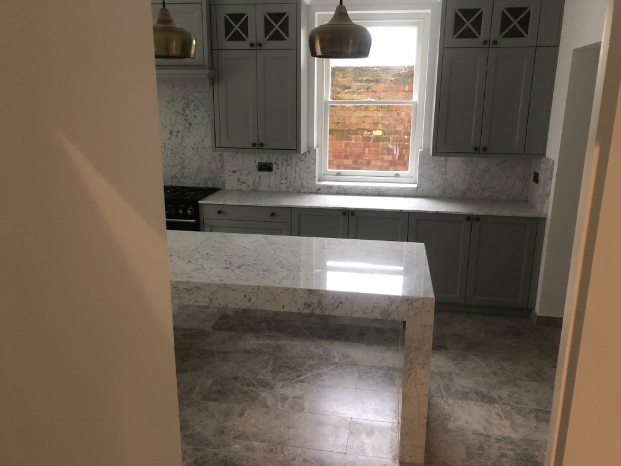 Nowoczesna kuchnia z szarymi szafkami, marmurowym blatem i podłogą, widok zza framugi drzwi.