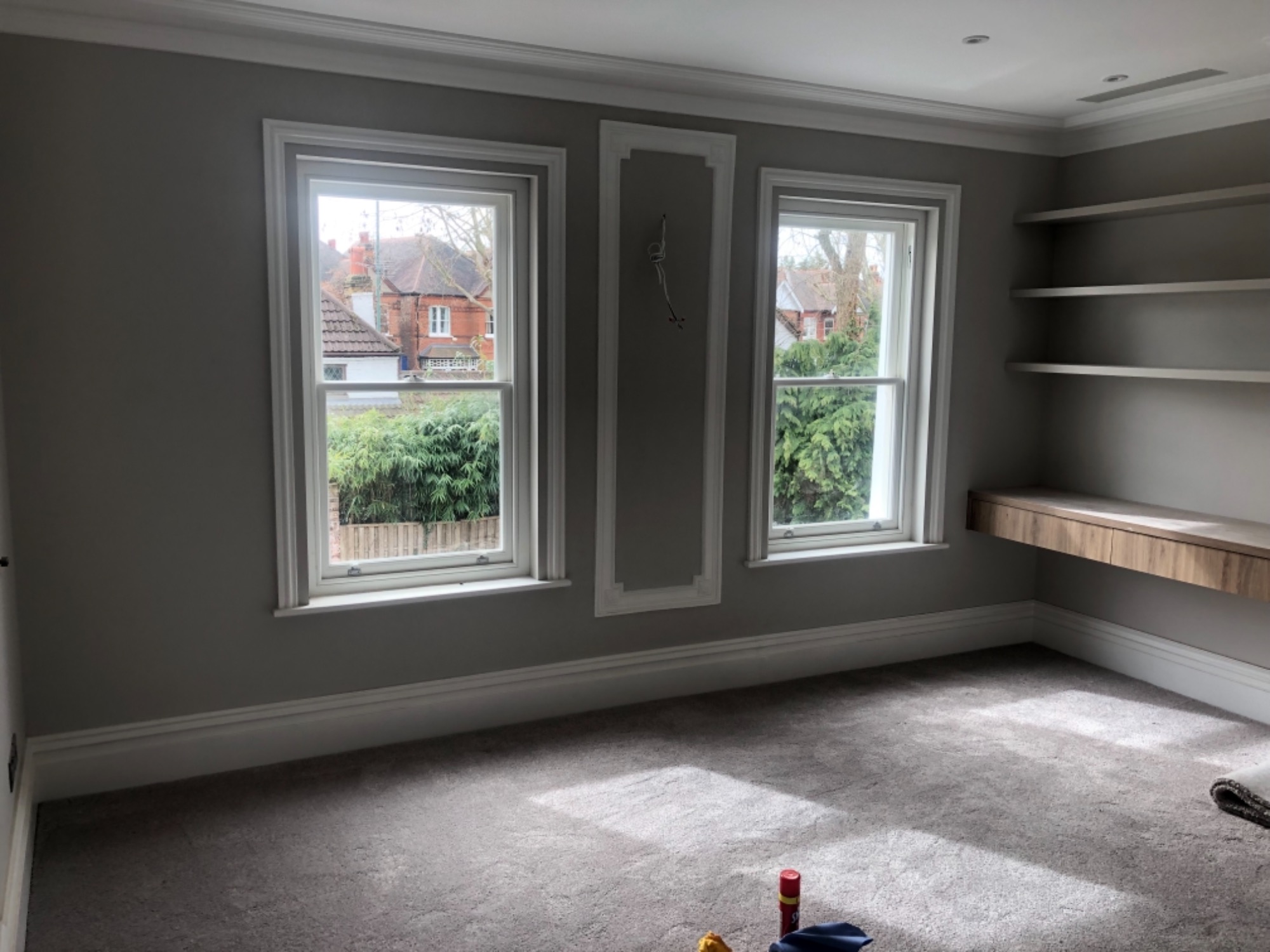 Pokój z dwoma oknami w białych ramach, szarymi ścianami z dekoracyjnymi listwami, szarą wykładziną i drewnianą półką. Widoczne narzędzia malarskie na podłodze sugerują świeżo zakończony remont.