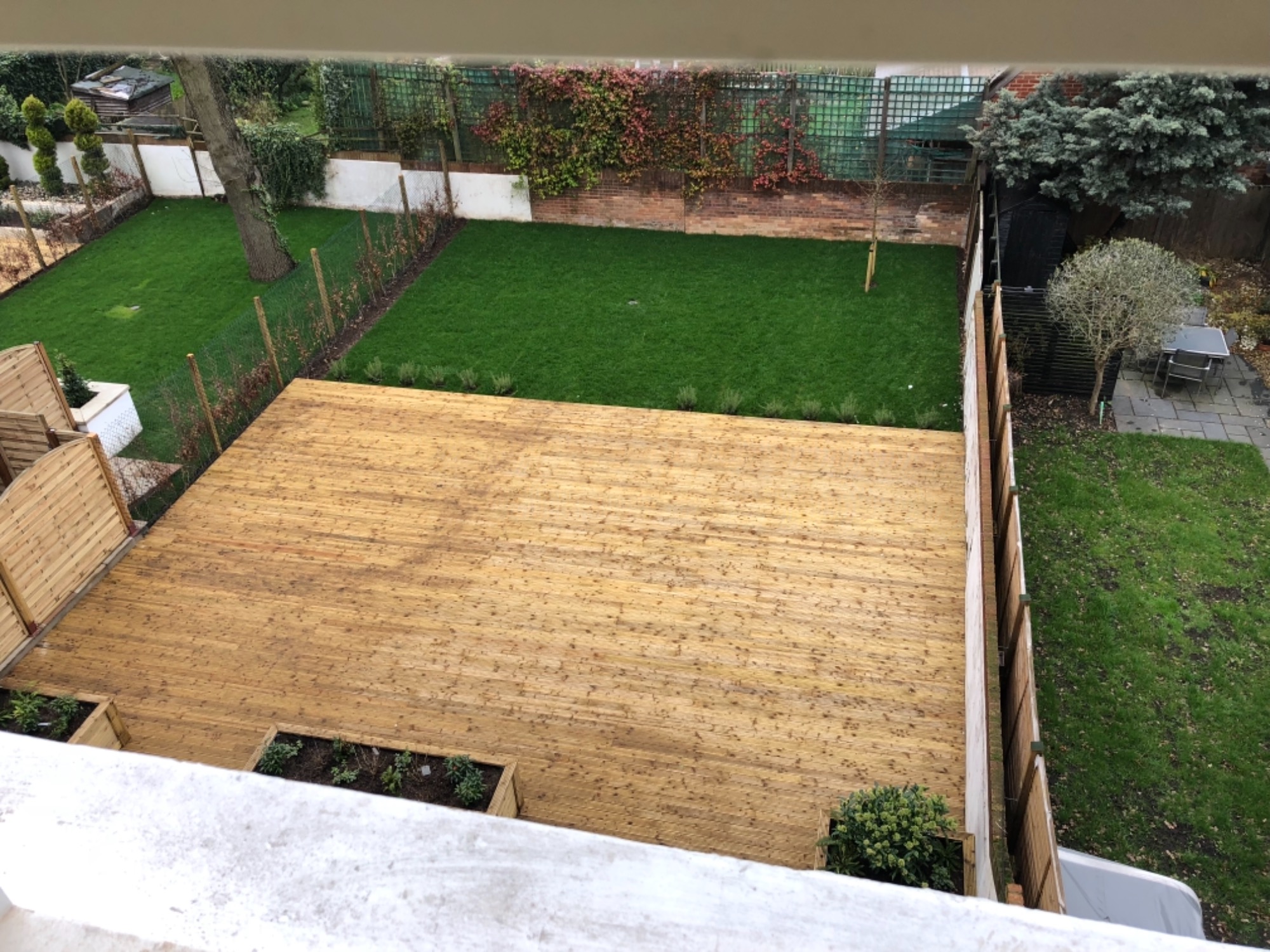Widok z góry na drewniany taras z wbudowanymi rabatami, przylegający do zadbanego trawnika z ozdobnymi nasadzeniami i ogrodzeniem.