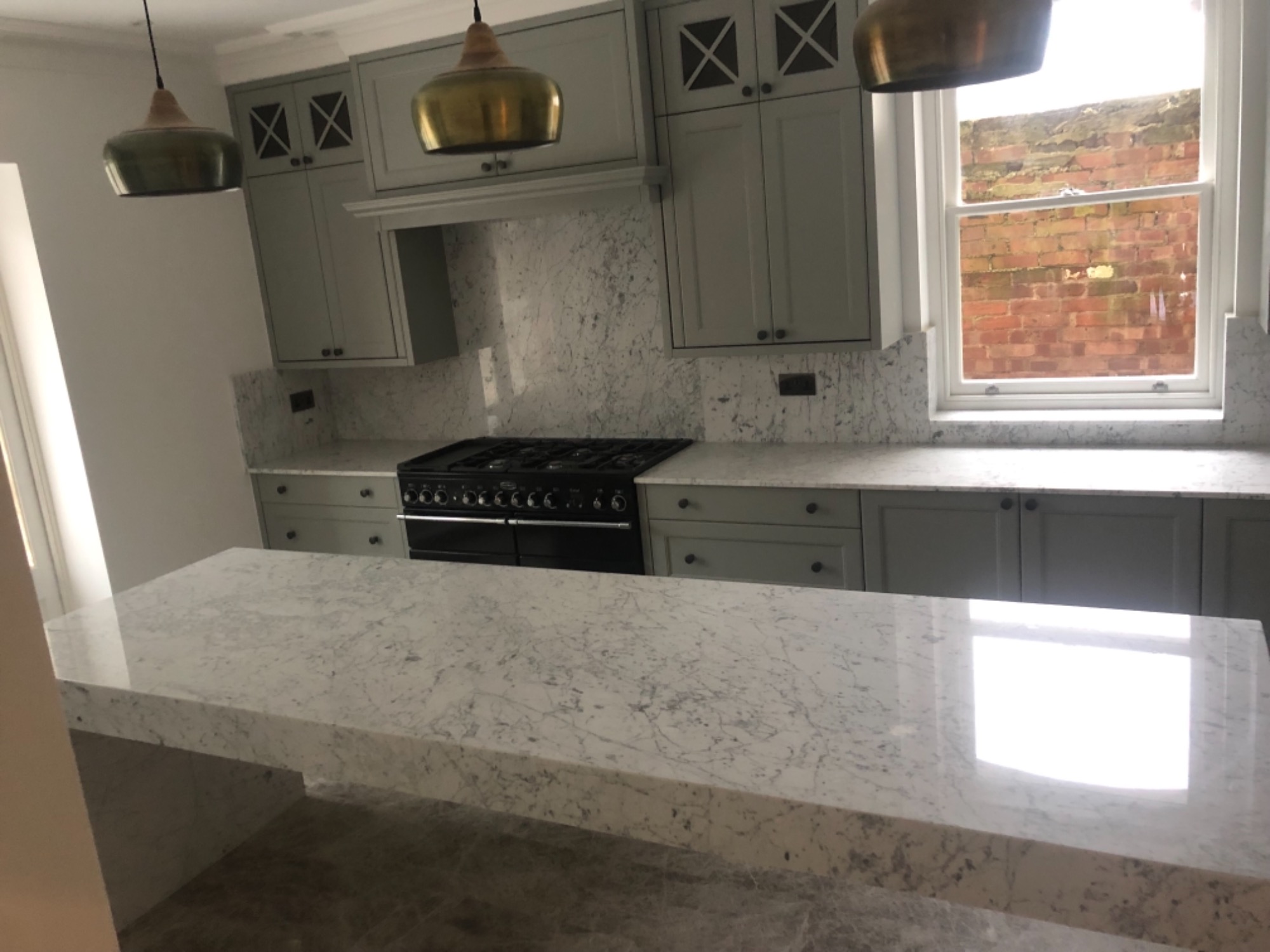 Wykończona kuchnia z szarymi szafkami, marmurowym blatem i złotymi lampami wiszącymi, widok na ceglany budynek przez okno.