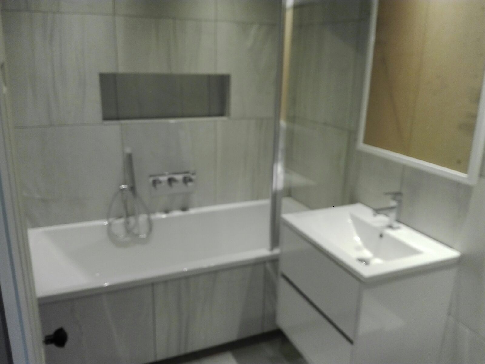 Nowoczesna łazienka z wanną obudowaną płytkami imitującymi marmur, bateria wannowa podtynkowa z trzema pokrętłami, umywalka nablatowa na szafce z dwiema szufladami i lustro w białej ramie.