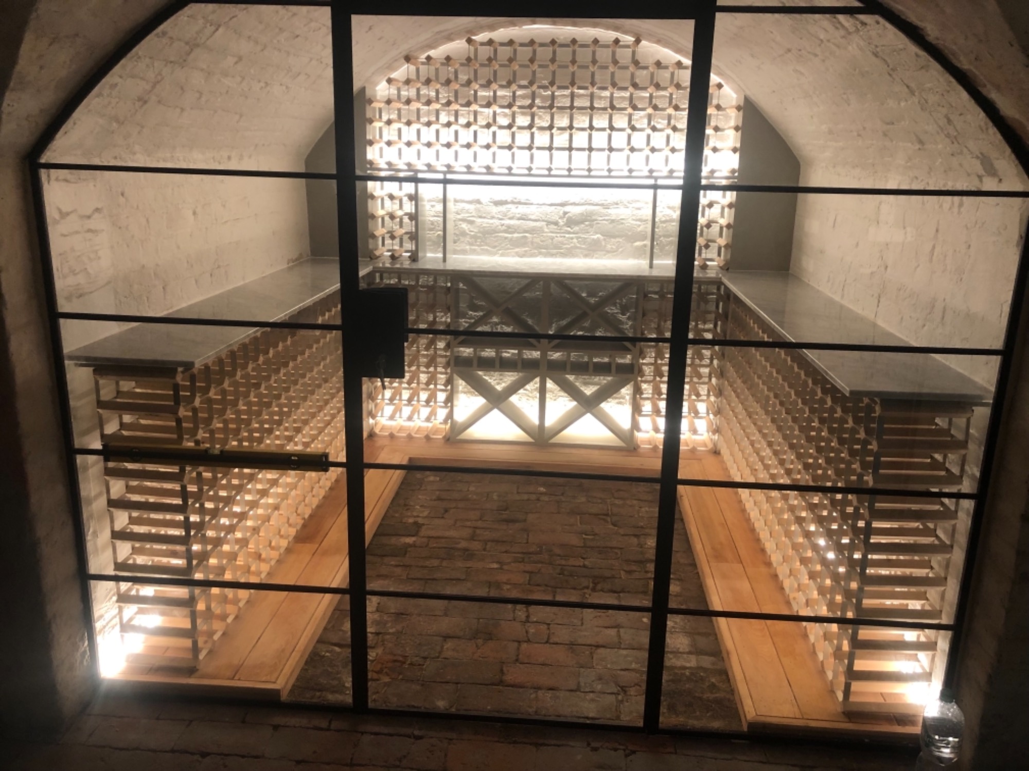 Piwnica z winami za czarną metalową kratą, ceglana podłoga, półki na wino z jasnego drewna, oświetlenie punktowe.