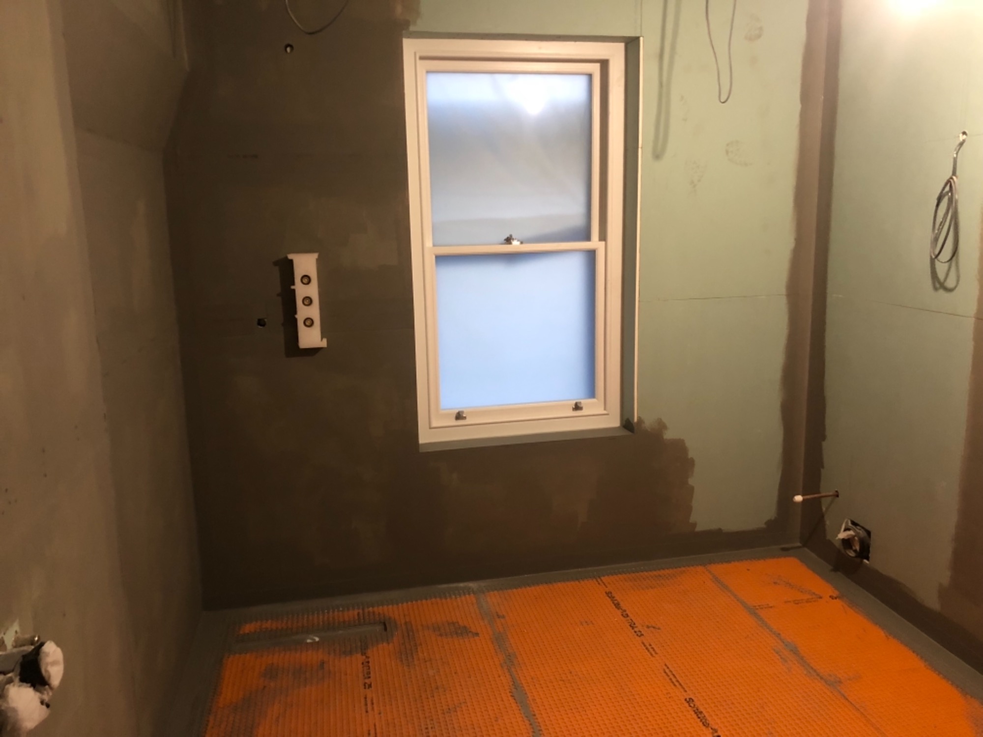 Przygotowanie łazienki do położenia płytek: ściany zagruntowane, na podłodze pomarańczowa mata izolacyjna, widoczne okno i instalacja pod baterię.