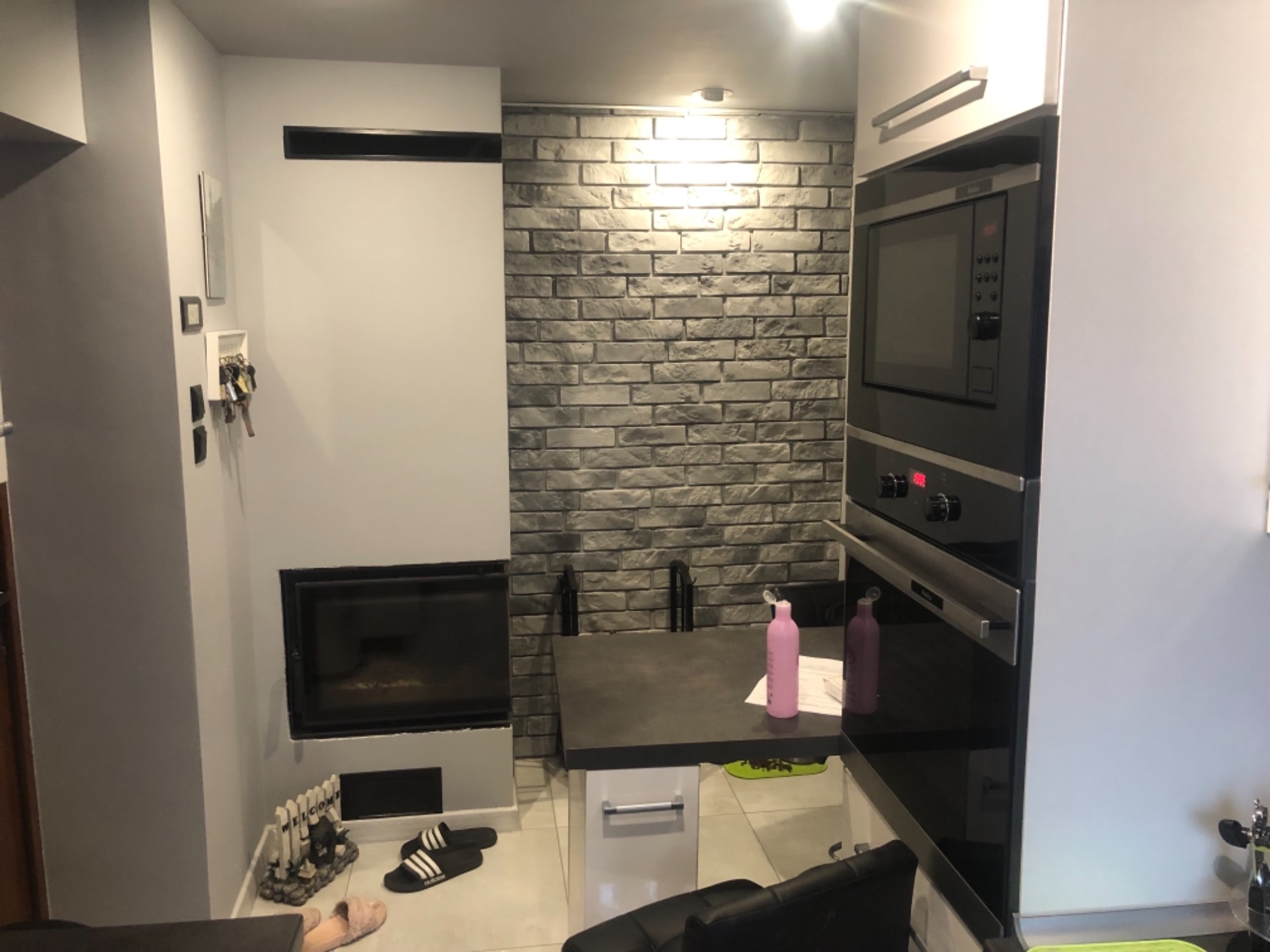 Nowoczesny salon z kominkiem obok kuchni, ściana z cegły dekoracyjnej, zabudowa kuchenna z piekarnikiem.