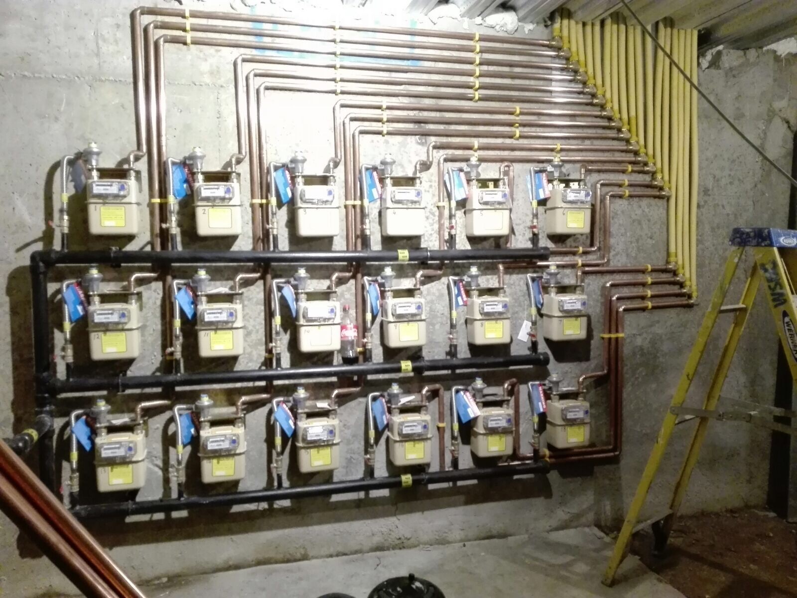 Rząd liczników gazowych zamontowanych na ścianie z betonowych płyt, z rurami miedzianymi i żółtymi rurami gazowymi, drabina w tle.