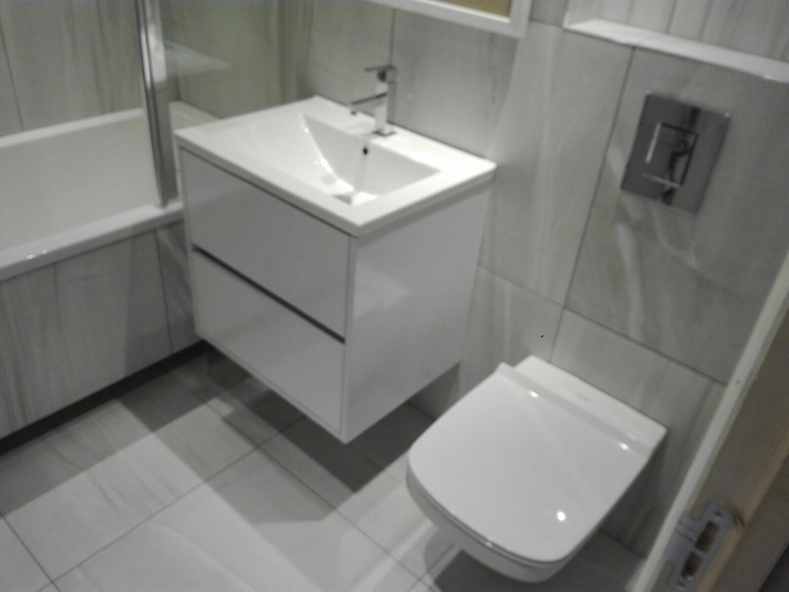 Nowoczesna, biała łazienka z podwieszaną szafką umywalkową, toaletą i wanną, wykończona szarymi płytkami imitującymi marmur.