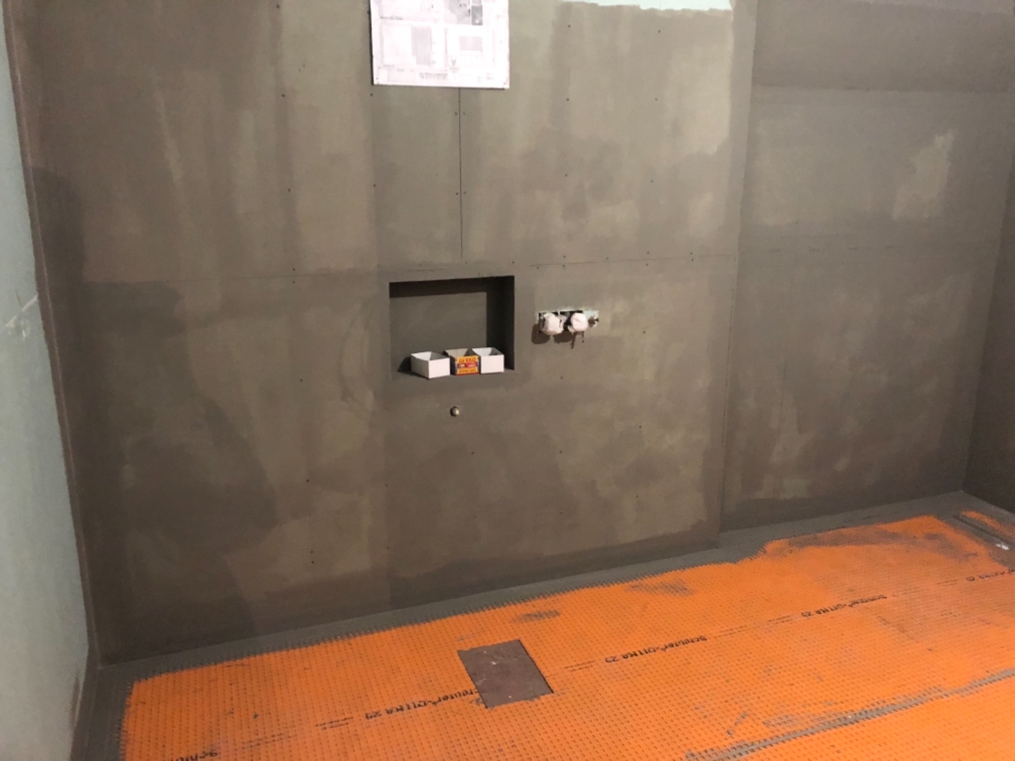 Przygotowana do wykończenia łazienka z pomarańczową matą grzewczą na podłodze, wnęką w ścianie i wyprowadzonymi instalacjami hydraulicznymi, na ścianie widoczny plan techniczny.