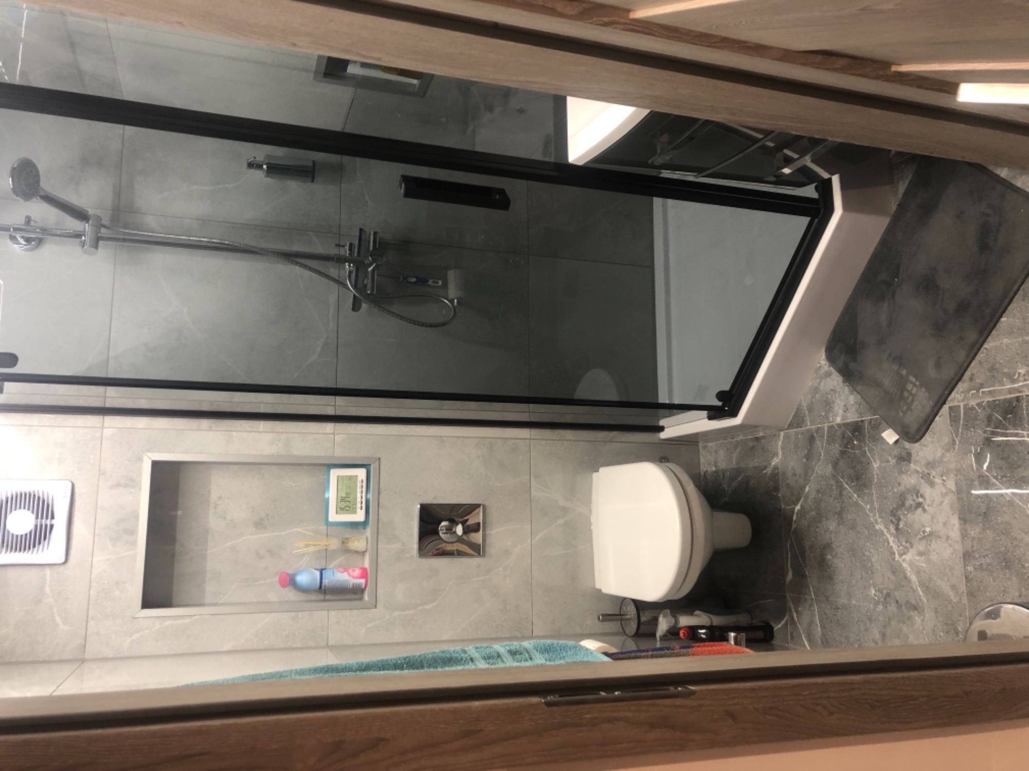 Nowoczesna łazienka z kabiną prysznicową z czarnymi ramami, toaletą, wnęką z półkami na kosmetyki i akcesoria, wykończona szarymi płytkami imitującymi marmur.