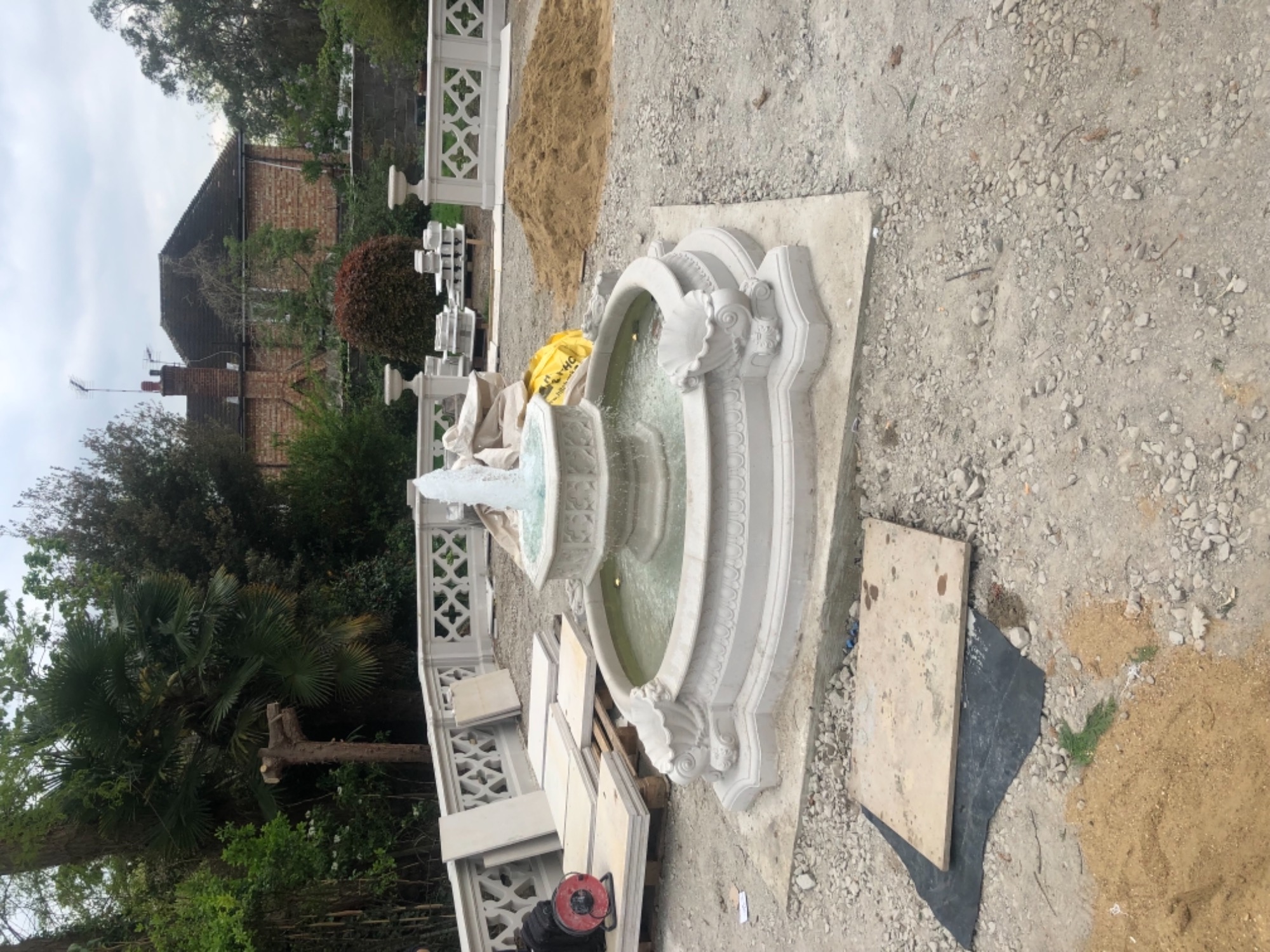 Fontanna ogrodowa z białego kamienia z tryskającą wodą, częściowo zmontowana, z elementami balustrady i stertą piasku w tle.