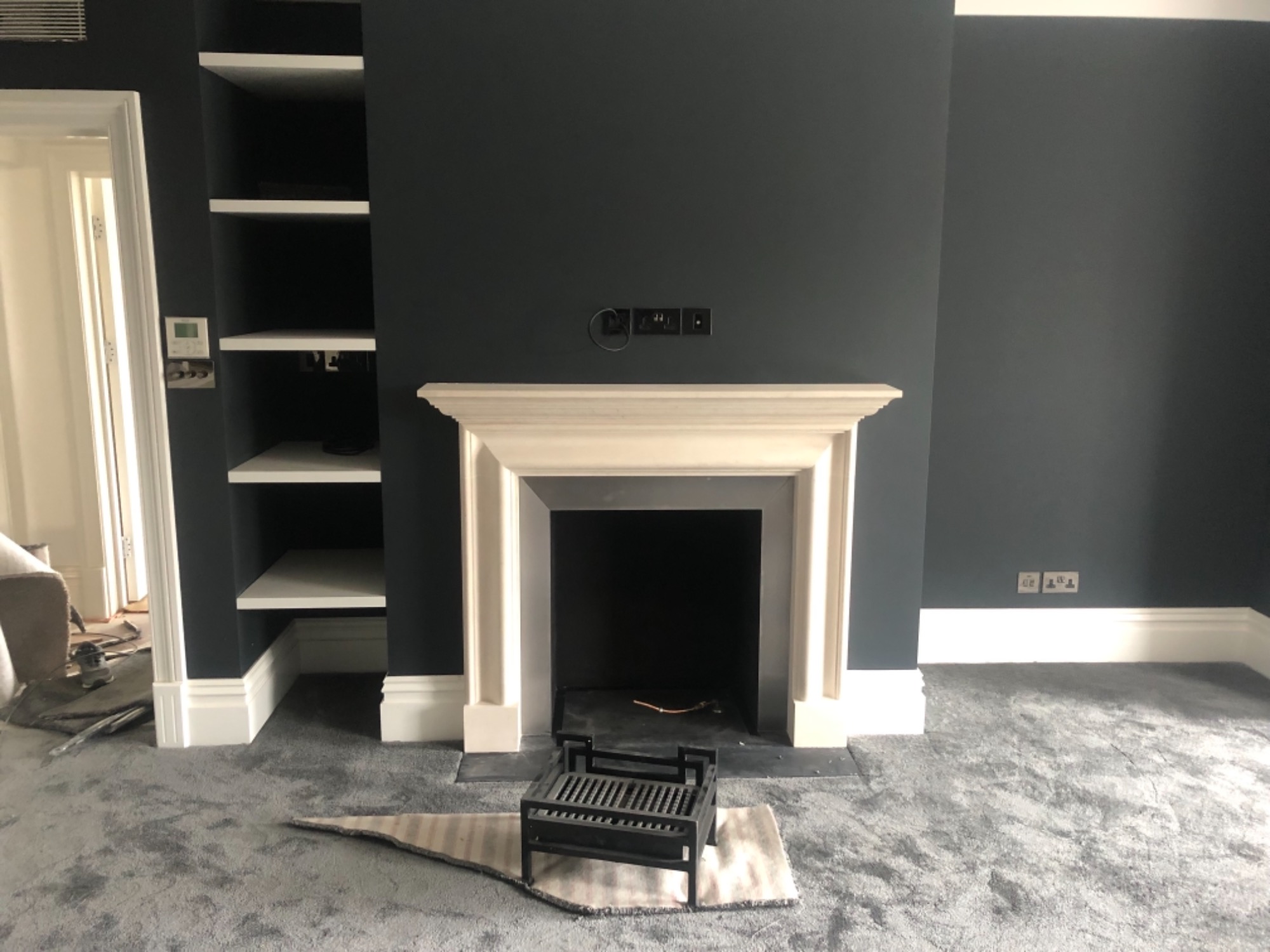 Pokój z ciemnymi ścianami, białym kominkiem i wbudowanymi półkami, widoczne gniazdka elektryczne i fragment niedokończonej podłogi.