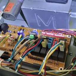 Wnętrze urządzenia AGD z widoczną płytą elektroniczną, licznymi przewodami w różnych kolorach, kondensatorem i elementami mocującymi, sugerujące proces naprawy lub diagnostyki.