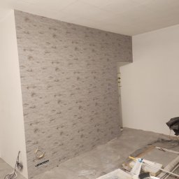 Usługi ogólno budowlane Sławomir gorzka. - Pokój w trakcie odświeżania: ściana pokryta wzorzystą tapetą na jednej ścianie, surowy beton na podłodze, widoczne kable elektryczne wystające ze ściany, narzędzia malarskie i folie ochronne...