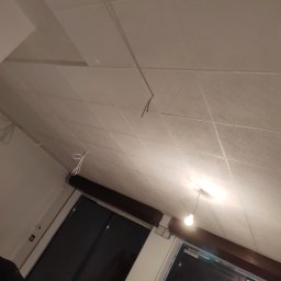 Usługi ogólno budowlane Sławomir gorzka. - Montaż sufitu podwieszanego kasetonowego z widocznymi przewodami oświetleniowymi, w tle okno zabezpieczone folią malarską.