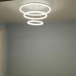 Usługi ogólno budowlane Sławomir gorzka. - Świeżo pomalowane na szaro wnętrze pokoju z oknem i nowoczesną lampą wiszącą składającą się z trzech okręgów LED