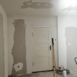 Usługi ogólno budowlane Sławomir gorzka. - Pokój w trakcie prac przygotowawczych do malowania: ściany z widocznymi śladami szpachlowania, zaklejone otwory, lampa budowlana na trójnogu oświetla wnętrze.