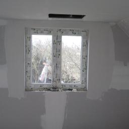 Usługi ogólno budowlane Sławomir gorzka. - Ściana w trakcie szpachlowania z widocznym oknem i kratką wentylacyjną pod sufitem. Ślady szpachli widoczne na ścianach, pianka montażowa wokół okna.