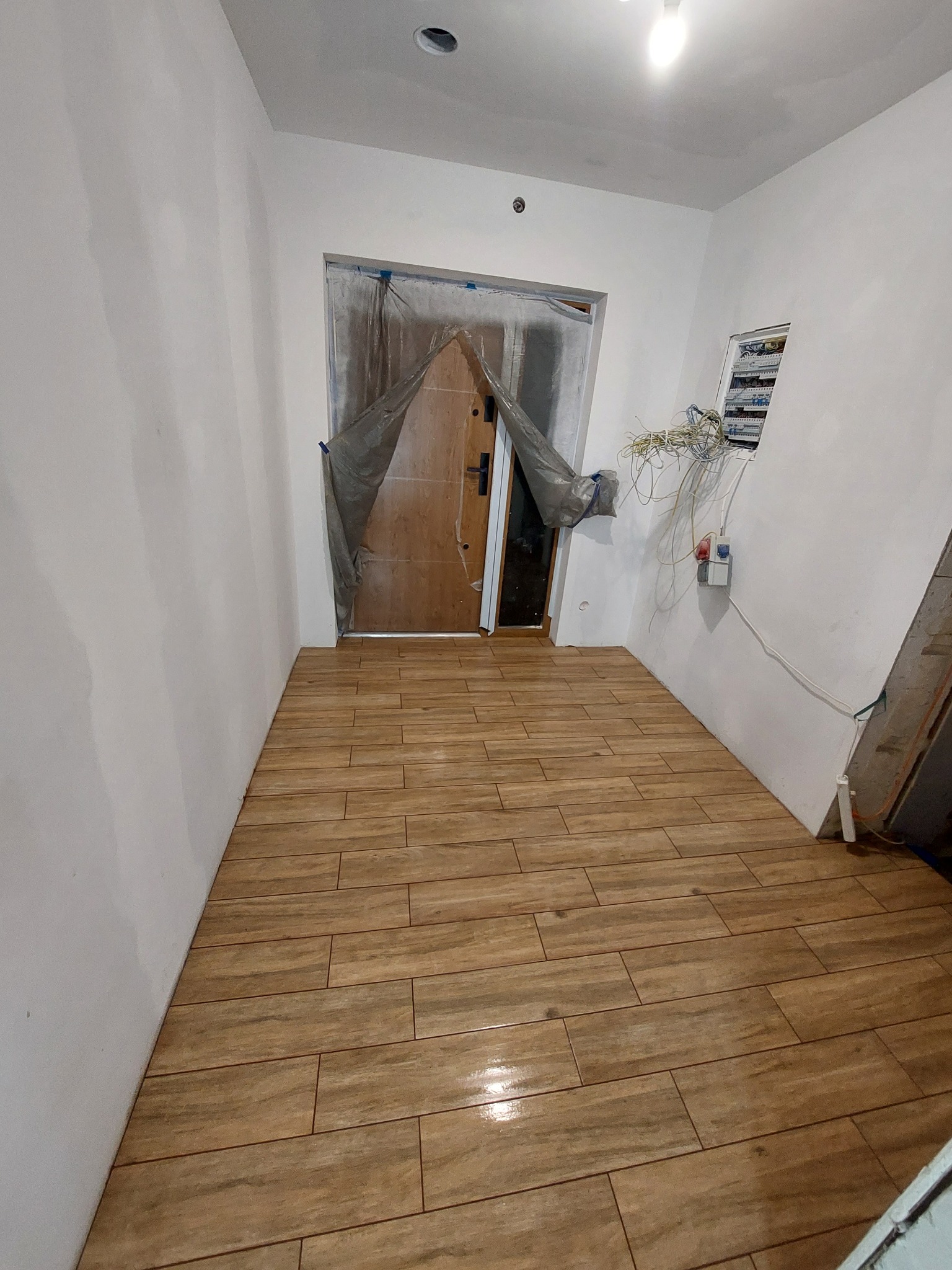 Wyłożona płytkami podłoga imitującymi drewno w korytarzu, zabezpieczone drzwi wejściowe folią malarską, widoczna otwarta skrzynka elektryczna z wystającymi przewodami.