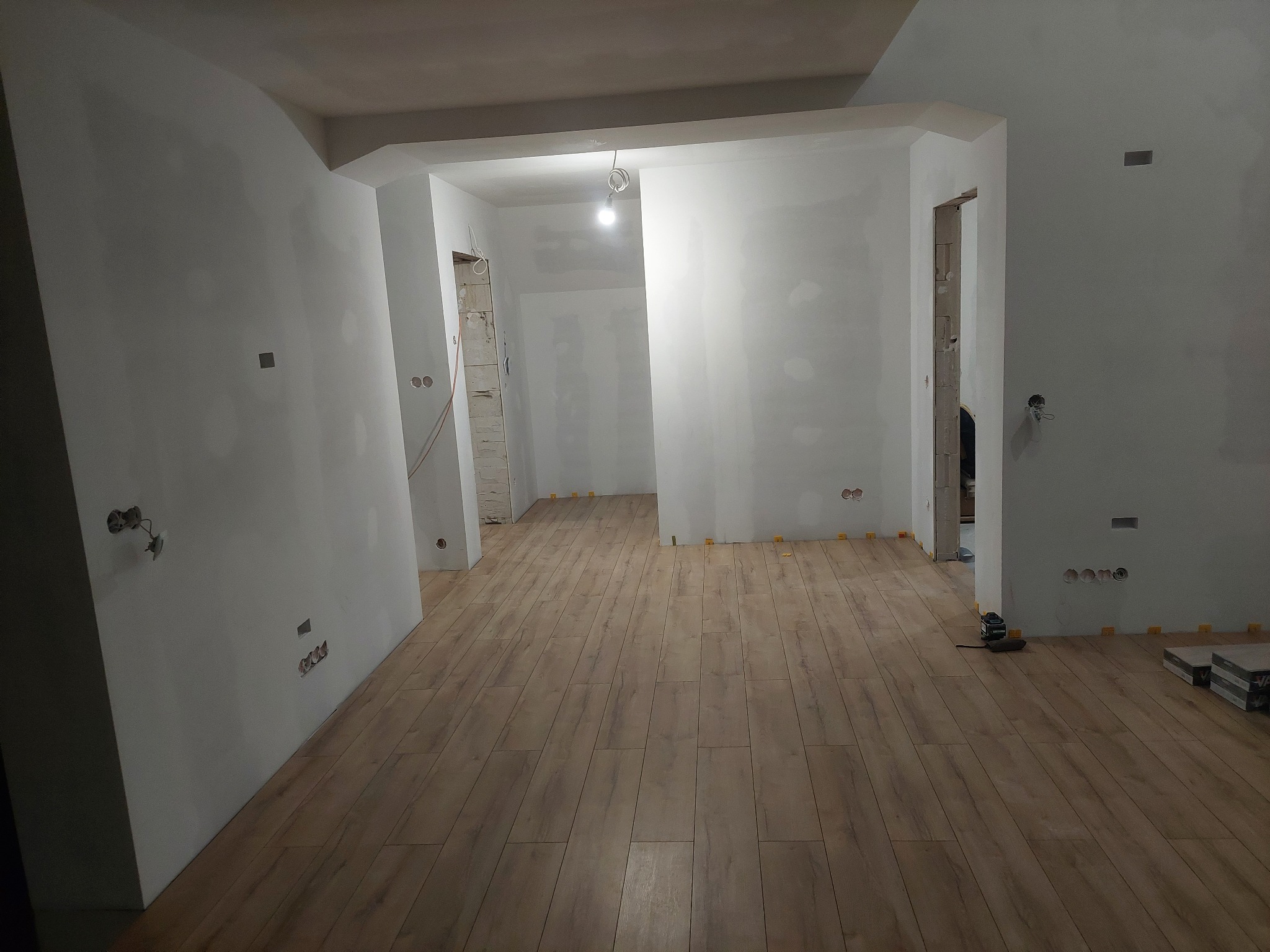 Układanie paneli podłogowych w jasnym, przestronnym wnętrzu w trakcie remontu, widoczne żółte kliny dystansowe i narzędzia pomiarowe.