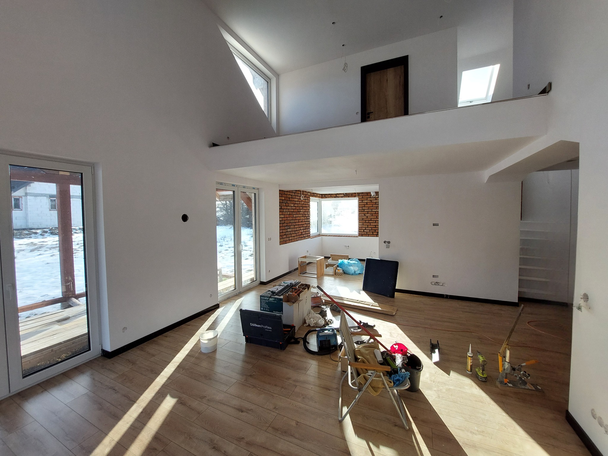 Wnętrze domu w trakcie wykańczania: białe ściany, drewniana podłoga, antresola z oknem dachowym, widoczne narzędzia i materiały budowlane w nieładzie, ceglana ściana w tle.